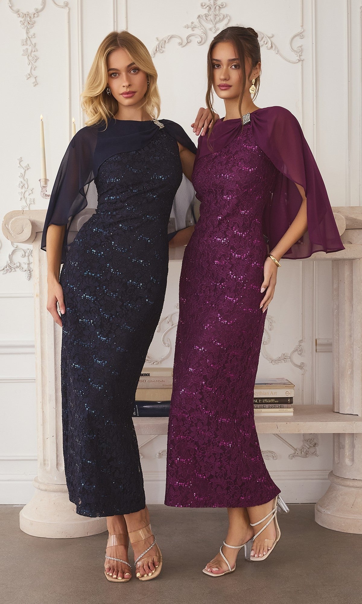 Two women stand side by side, posing indoors against an ornate white wall. One wears the Amoris T906 Long Prom Dress in eggplant with a sequin finish and chiffon cape overlay; the other wears a similar navy blue gown.